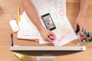 A person working on a podcast business plan, highlighting key ideas on paper with a pink marker. The desk is covered with brainstorming sketches, a smartphone, and various office supplies, illustrating the planning and strategy behind podcast development.