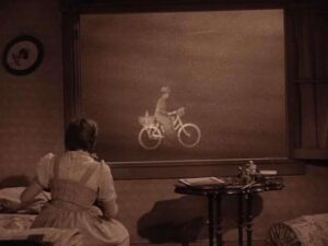 A woman watches a bicyclist pass outside a window during a surreal tornado scene.