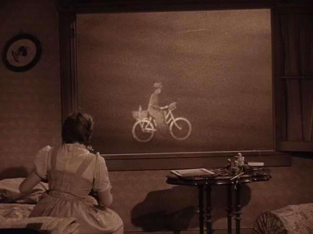 A woman watches a bicyclist pass outside a window during a surreal tornado scene.